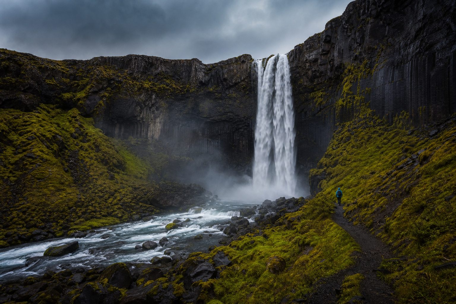 A powerful waterfall over dark basalt cliffs and green moss in remote Iceland, a tiny hiker for scale
